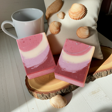 Red Feathers Organic Artisan Soap bars on a wooden surface with seashells and a white mug in the background.