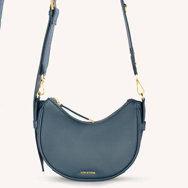 Mira & Moss Sabine Crescent Crossbody Bag in blue vegan leather with gold-tone hardware and adjustable strap.