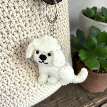Bichon Puppy Dog Felt Wool Keychain hanging from a textured cream bag